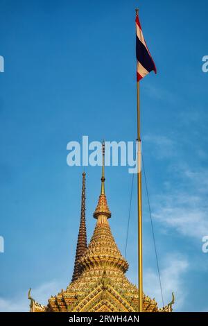 Ein thailändischer Tempelturm und eine Flagge vor einem blauen Himmel. Der Tempelturm ist reich verziert und hat eine goldene Farbe. Die Flagge ist rot, weiß und blau und fliegt weiter Stockfoto