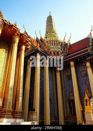 Ein thailändischer Tempel mit verzierten goldenen und blauen Säulen, einem hohen Turm und einem roten Dach mit goldenen Akzenten sonnt sich in einem Innenhof. Stockfoto