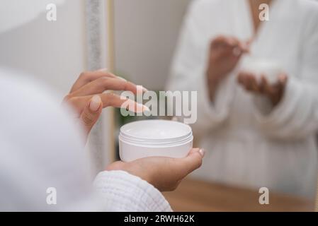 Frau, die Hände hält Lotionglas am Frisiertisch im Badezimmerkleid, die Anti-Aging-Produkt nimmt Stockfoto