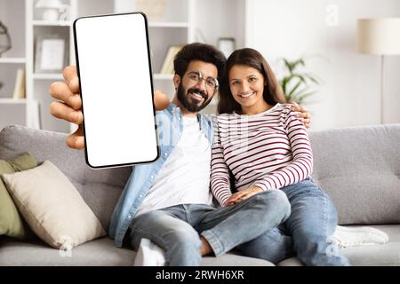 Nettes, glückliches indisches Paar, das auf dem Sofa sitzt, Telefon zeigt, Mockup Stockfoto