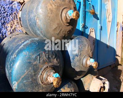 Ein Stapel von Gasflaschen aus verstörtem blauem Metall liegt an ihren Seiten übereinander, vor einer rauen strukturierten blauen Wand und gemusterten Türen. Stockfoto