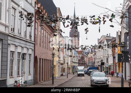 Flensburg, Deutschland - 10. Februar 2017: Sportliche Schuhe hängen an Drähten. Gewöhnliche Leute gehen die Straße der alten Flensburger Stadt entlang Stockfoto