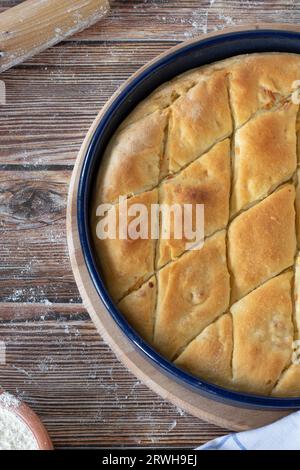 Frisch gebackener Kuchen in einer Pfanne auf Holzuntergrund, mit Mehl und Stecknadel bestäubt. Draufsicht, vertikale Aufnahme. Traditionelles Balkan-Essen. Stockfoto