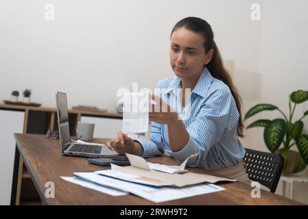 Die junge Dame sitzt am Tisch und berechnet die Zahlungen der Versorgungsunternehmen, bevor sie in der Online-Bank-App Geld überweist Stockfoto