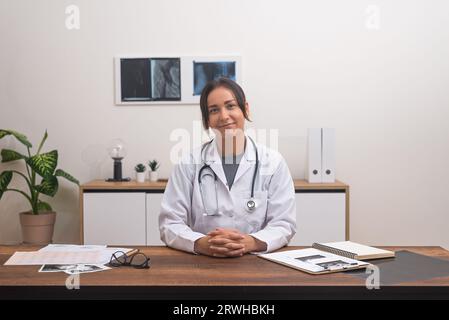 Junge Ärztin im Labormantel lächelt am Schreibtisch mit Patientendokumenten und schaut in die Kamera Stockfoto