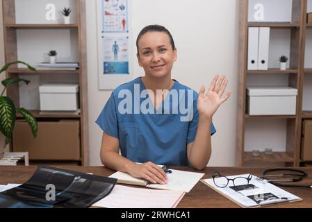 Lächelnde Ärztin im blauen Labormantel winkt Hand in Gruß an den Patienten, der die Kamera anschaut Stockfoto