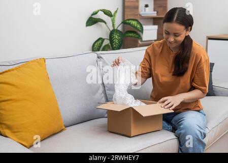 Eine fröhliche Käuferin nimmt Luftpolsterfolie aus einer kleinen Schachtel mit Waren heraus, die auf dem Sofa im Wohnzimmer sitzen Stockfoto