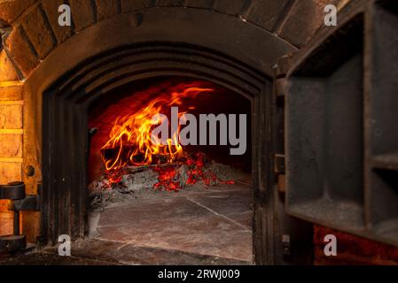 Traditioneller Brennholzofen mit Holzbrand mit Flammen und brennender Glut von gusseiserner Tür an der Backsteinwand aus gesehen Stockfoto