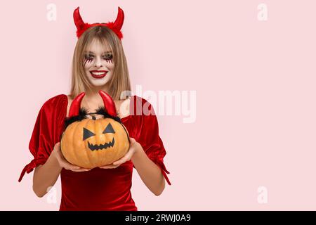 Junge Frau, die zu Halloween als Teufel mit Kürbis auf rosafarbenem Hintergrund gekleidet ist Stockfoto