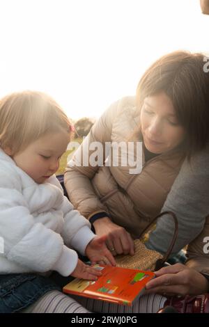 Vor herbstlicher landschaftlicher Kulisse teilen sich eine Mutter und ihre blonde Einjahres-Tochter einen Moment, der in einem illustrierten Buch vertieft ist. Sie sind in Warml gekleidet Stockfoto