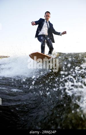 Ein junger Mann im klassischen Anzug fährt auf einem Wakeboard auf dem Fluss oder See in der Nähe der Stadt. Ein unvorsichtiger Mitarbeiter entkam aus einem stickigen Büro, um seine Lieblingsakti aufzunehmen Stockfoto
