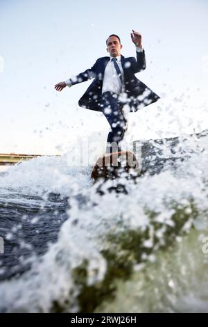 Ein junger Mann im klassischen Anzug fährt auf einem Wakeboard auf dem Fluss oder See in der Nähe der Stadt. Ein unvorsichtiger Mitarbeiter entkam aus einem stickigen Büro, um seine Lieblingsakti aufzunehmen Stockfoto
