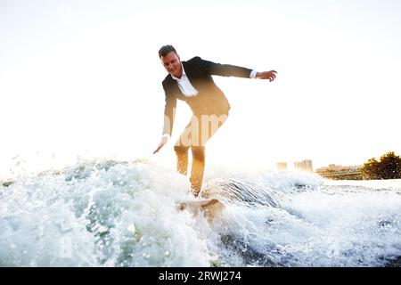 Ein junger Mann im klassischen Anzug fährt auf einem Wakeboard auf dem Fluss oder See in der Nähe der Stadt. Clerk entkam aus einem stickigen Büro, um seinen Lieblingssport aufzunehmen. B Stockfoto