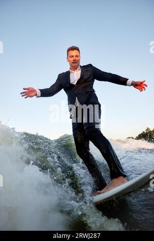 Ein junger Mann im klassischen Anzug fährt auf einem Wakeboard auf dem Fluss oder See in der Nähe der Stadt. Ein unvorsichtiger Mitarbeiter entkam aus einem stickigen Büro, um seine Lieblingsakti aufzunehmen Stockfoto