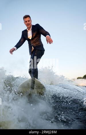 Ein junger Mann im klassischen Anzug fährt auf einem Wakeboard auf dem Fluss oder See in der Nähe der Stadt. Ein unvorsichtiger Mitarbeiter entkam aus einem stickigen Büro, um seine Lieblingsakti aufzunehmen Stockfoto