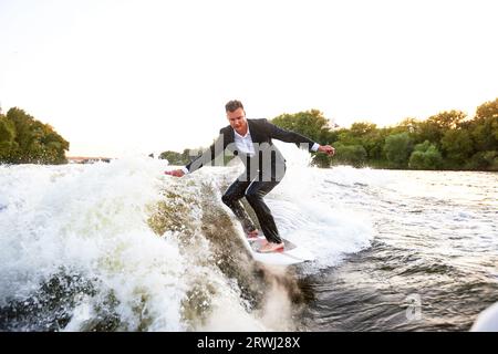Ein junger Mann im klassischen Anzug fährt auf einem Wakeboard auf dem Fluss oder See in der Nähe der Stadt. Ein unvorsichtiger Mitarbeiter entkam aus einem stickigen Büro, um seine Lieblingsakti aufzunehmen Stockfoto