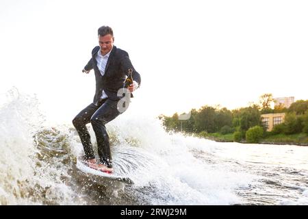 Junger Mann im klassischen Anzug mit Champagnerflasche fährt auf einem Wakeboard auf dem See in der Nähe der Stadt. Happy Clerk ist aus einem stickigen Büro geflohen, um aktive sp Stockfoto
