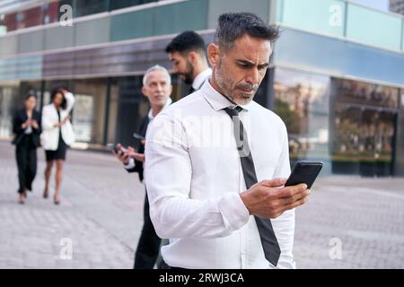 Ein Geschäftsmann mittleren Alters sieht sich ein Handy an, das sich konzentriert und in einer Menschenmenge die Straße entlang läuft. Stockfoto