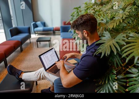 Ein männlicher Entwicklungsingenieur, der an einem Projekt arbeitet, verwendet einen Laptop, während er in gemütlicher Coworking-Umgebung sitzt Stockfoto