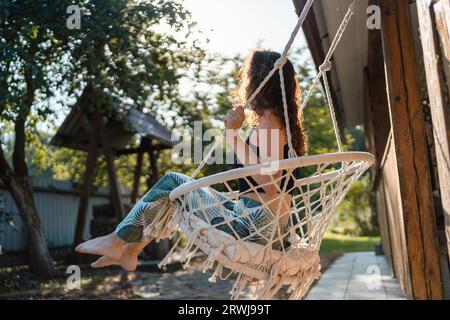 Glückliche junge Frau, die auf einem Makrame-Schaukelstuhl in der Nähe des Landhauses im Freien sitzt Stockfoto