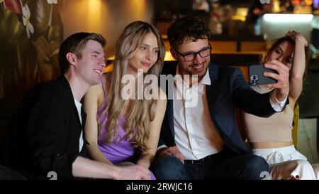 Lächelnde junge Freunde sitzen im Restaurant, während sie Fotos machen, mit dem Smartphone fotografieren und sich vor der Kamera positionieren. Lifestyle- und Freudenkonzept. Zeitlupe Stockfoto