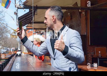 Kaukasischer, erwachsener Geschäftsmann, der vor dem Restaurant auf einer Holztheke sitzt und auf sein Telefon schaut, während er auf eine Kellnerin wartet Stockfoto