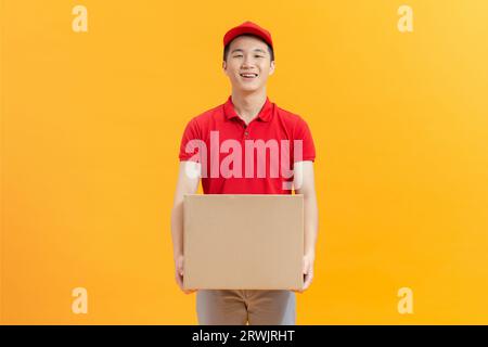 Freundlicher asiatischer Liefermann trägt rotes Hemd und Kappe mit Paketpaket auf gelbem Hintergrund Stockfoto