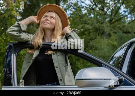 Blonde Frau mit Hut, die neben der Autotür wohnt. Junge Touristen erkunden lokale Reisen und machen echte Momente. Wahre Emotionen Ausdruck des Ausgehens und der Erfrischung Entspannen Sie sich an der frischen Luft Stockfoto