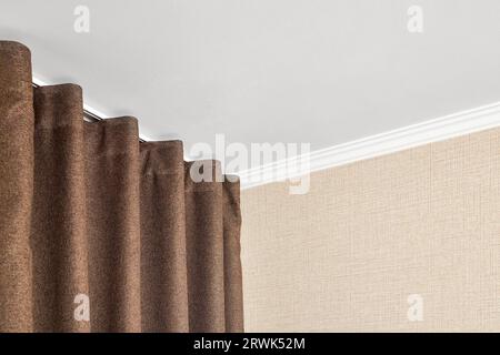 Vorhänge aus braunem Mattenstoff hängen am Deckengesims in der Nähe der Wand, die mit einer Mattenstruktur oder einem rauen Canvas unter einem weiß tapeziert sind Stockfoto