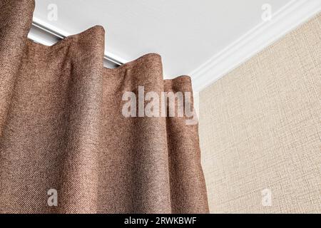 Vorhänge aus braunem Mattenstoff hängen am Deckengesims in der Nähe der Wand, die mit einer Mattenstruktur oder einem rauen Canvas unter einem weiß tapeziert sind Stockfoto