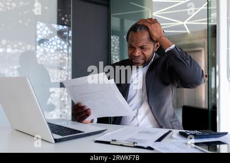 Frustrierter und verärgerter Finanzier am Arbeitsplatz im Büro, verärgerter Chef, der Rechnungen, Verträge und Berichte prüft, mit Laptop am Tisch sitzt, Geschäftsmann hinter Papierarbeit. Stockfoto