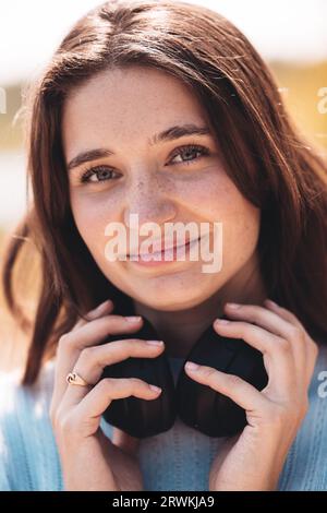 In diesem fesselnden Außenporträt steht eine junge Millennials-Frau selbstbewusst in einem Stadtpark und strahlt natürliche Schönheit und Selbstvertrauen aus. Sie trägt ein weiches blaues Hemd und hält ihre Kopfhörer um den Hals, um ihren sorglosen Geist zu zeigen. Diese hübsche junge Frau verkörpert das Wesen der Selbstsicherheit und den Reiz der freien Natur. Selbstbewusste Millennials im Park: Eine junge Frau, die sich der natürlichen Schönheit verschrieben hat. Hochwertige Fotos Stockfoto