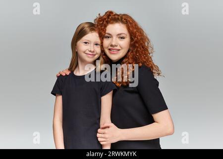 Zwei-Generationen-Konzept, fröhliche rothaarige Mutter und Tochter in passender Kleidung, die sich auf Grau umarmt Stockfoto