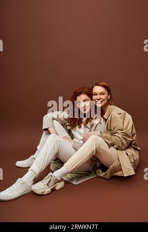 Zwei Generationen, positive Frauen mit rotem Haar, die in Herbsttrenchmänteln auf braunem Hintergrund posieren Stockfoto