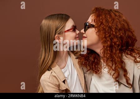 Zwei Generationen, glückliche rothaarige Mutter und Tochter in Sonnenbrille auf braunem Hintergrund, von Angesicht zu Angesicht Stockfoto