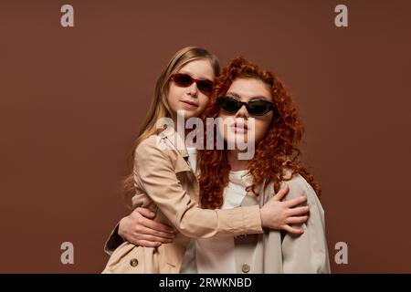 Zwei Generationen, rothaarige Mutter und Tochter in Sonnenbrille und beigefarbene Trenchcoat auf braunem Hintergrund Stockfoto