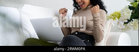 Erfolgreiche afroamerikanische Geschäftsfrau fühlt sich glücklich über die entscheidende Arbeit am Laptop, während sie zu Hause auf der Sofaecke sitzt Stockfoto