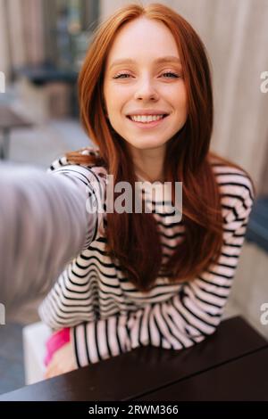 Vertikale POV-Aufnahme einer glücklichen jungen Frau, die Videoanrufe redet oder Selfie-Bilder macht, die Kamera am Tisch im Café im Freien betrachten. Stockfoto