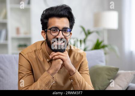 Nahaufnahme eines jungen lächelnden lateinamerikanischen Mannes, der lächelt und in die Kamera schaut, während er im Wohnzimmer auf dem Sofa sitzt. Stockfoto