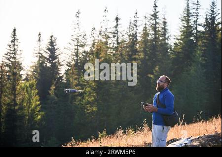 Professioneller Fotograf, der Fotos von der Drohne macht. Junger Mann, der Luftaufnahmen in den Bergen macht. Mann, der seine Drohne auf dem Hintergrund des Bergwaldes kontrolliert. Stockfoto