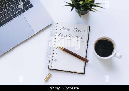 Öffnen Sie den Notizblock mit Text für Neujahrsvorsätze. Und leere Liste. Blume, Laptop und Kaffeetasse im Hintergrund. Stockfoto
