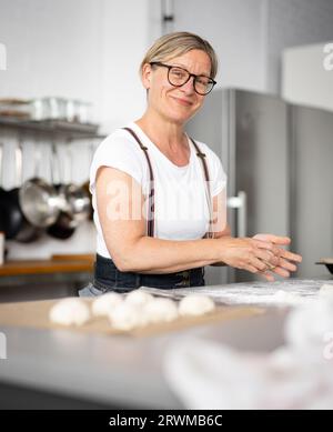 Eine Frau steht in einer schönen Küche, bereit zum Backen. Sie knetscht Teig für die Herstellung von Brötchen und schaut mit einem fröhlichen Lächeln in die Kamera. Stockfoto