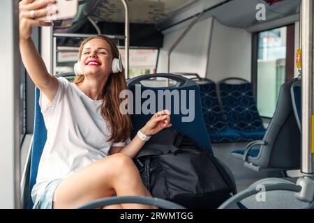 Unvorsichtige junge Frau mit weißem T-Shirt, rotem Lippenstift und Kopfhörern nimmt ein Selfie, während sie im öffentlichen Shuttlebus mit ihrem Mobiltelefon sitzt. Stockfoto