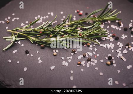 Ein Blumenstrauß aus Rosmarin auf dem Tisch auf einem schwarzen Granitteller, verstreutes Himalaya-Rosa-Salz und schwarze Pfefferkörner Stockfoto