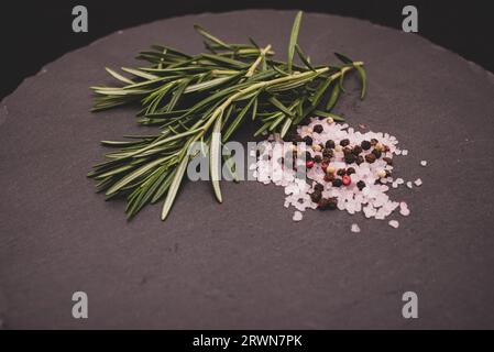 Ein Blumenstrauß aus Rosmarin auf dem Tisch auf einem schwarzen Granitteller, verstreutes Himalaya-Rosa-Salz und schwarze Pfefferkörner Stockfoto