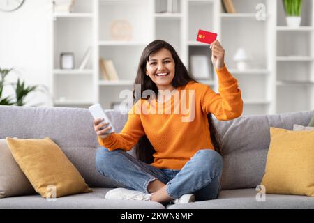Fröhliche junge indianerin, die zu Hause Telefon und Kreditkarte benutzt Stockfoto