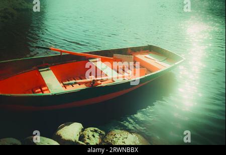 Ein altes rotes Fischerboot mit Rudern schwimmt an einem ruhigen Sommertag am Flussufer und spiegelt die hellen Sonnenstrahlen auf seiner Oberfläche wider. Die Nostalgie, Leisurel Stockfoto