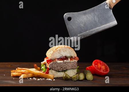 Hausgemachter Hamburger in der Mitte mit Rindfleisch, Tomaten, Salat, Käse und Pommes Frites auf Holztisch. Fleischspeck in der Hand. Fastfood auf dunklem BA Stockfoto