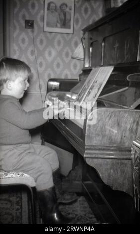 1960er Jahre, historisch, in einem Vorderzimmer, ein kleiner Junge, der Klavier spielt... und seine wellington-Stiefel trägt! Stockfoto