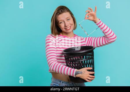 Attraktive Frau in Crop Top, die nach einer medizinischen Vision-Lasertherapie-Operation, die lächelnd auf die Kamera schaut, die Brille in den Abfalleimer wirft. Gesundes kuriertes Mädchen isoliert auf blauem Hintergrund Stockfoto